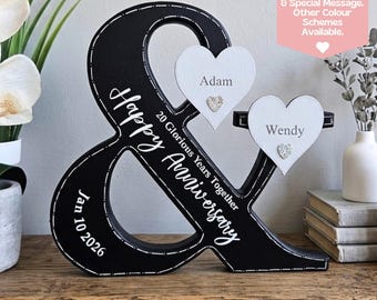 Puede incluir: Decoración de aniversario de madera negra en forma de ampersand con texto blanco que dice "Happy Anniversary" y "20 Glorious Years Together". Se adjuntan dos placas blancas en forma de corazón, cada una con un nombre y una gema. También se muestra la fecha "Jan 10 2026".