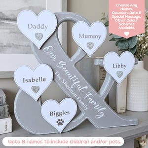 May include: A grey wooden ampersand sign featuring five white heart-shaped plaques with the names Daddy, Mummy, Isabelle, Libby, and Biggles. The text "Our Beautiful Family" and "The Sherman Family" is printed on the ampersand with small silver hearts.