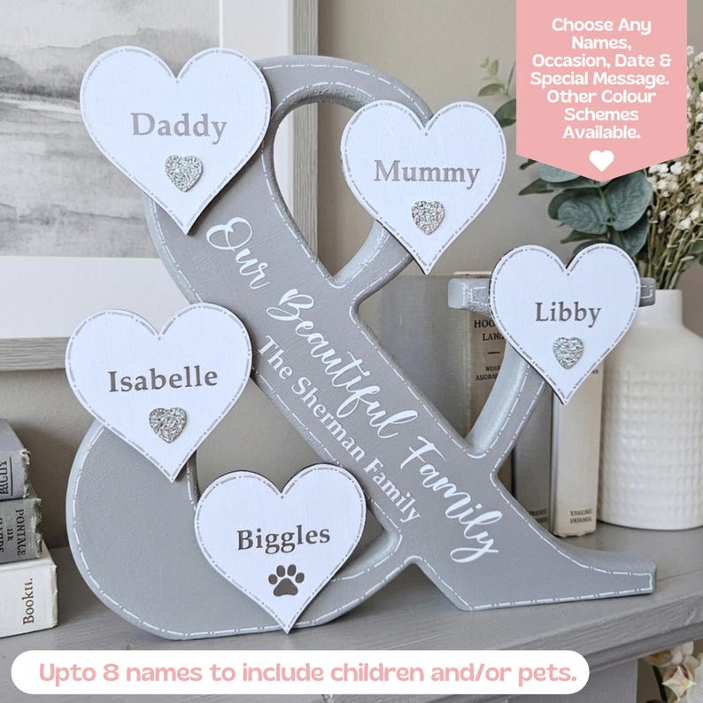 May include: A grey wooden ampersand sign featuring five white heart-shaped plaques with the names Daddy, Mummy, Isabelle, Libby, and Biggles. The text "Our Beautiful Family" and "The Sherman Family" is printed on the ampersand with small silver hearts.