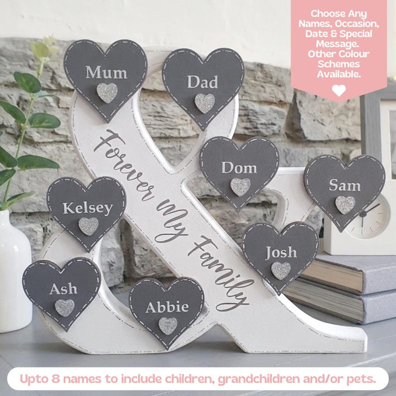 May include: A white wooden ampersand sign featuring the text Forever My Family, decorated with seven grey heart-shaped plaques. Each heart displays a name and a small silver glitter heart accent. The sign includes a pink banner with text.