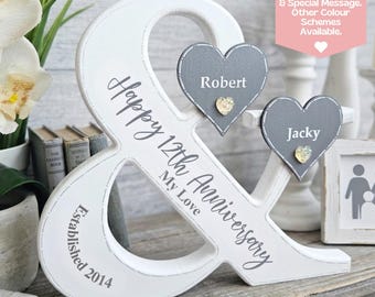 Puede incluir: Un letrero de madera blanco con forma de ampersand que presenta el texto "Happy 12th Anniversary MY LOVE" y "Established 2014" en gris. Se incluyen dos placas de madera grises en forma de corazón con los nombres "Robert" y "Jacky" y detalles dorados.