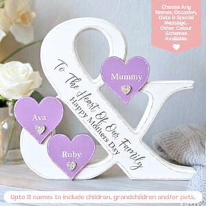 May include: A white wooden ampersand sign featuring the text To the Heart of Our Family Happy Mothers Day. Three lavender heart-shaped plaques are attached, each displaying a name: Mummy, Ava, and Ruby, with a small silver heart accent.