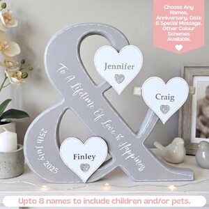 May include: A grey ampersand-shaped wooden sign featuring the white text To A Lifetime Of Love & Happiness and 25th July 2025. Three white heart-shaped plaques are attached, displaying the names Jennifer, Craig, and Finley with small silver glitter hearts.