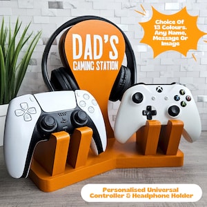 May include: Orange wooden stand with two white game controllers and a black headset. The stand has the words "DAD'S GAMING STATION" on it.