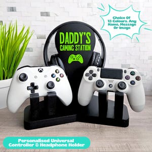 May include: A black headphone stand with the text "DADDY'S GAMING STATION" in green, holding a pair of headphones. Two white game controllers rest on black stands. The image also includes the text "Personalised Universal Controller & Headphone Holder".