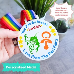 May include: A white personalised medal with a rainbow ribbon. The medal features a green fairy with wings, a dummy, and the text "You Are So Brave Lochlan Love From The Paci Fairy".