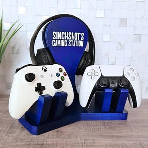 May include: A blue gaming station with the text "SINGHSHOT'S GAMING STATION" in white. The station holds a pair of black headphones, a white Xbox controller, and a white PlayStation 5 controller.