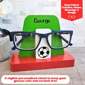 May include: A green and red wooden glasses stand with a soccer ball design. The stand has a slot for glasses and a space for a name, in this case, "George".