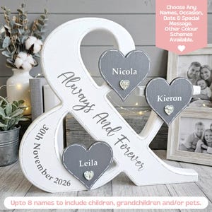 May include: A white wooden ampersand sign featuring the text Always And Forever and 30th November 2026. Three grey wooden hearts are attached, each displaying a name: Nicola, Kieron, and Leila, with a small clear crystal accent on each heart.