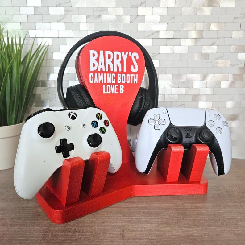May include: A red wooden gaming station with the text "BARRY'S GAMING BOOTH LOVE B". It holds a pair of black headphones, a white Xbox controller, and a white PlayStation controller.