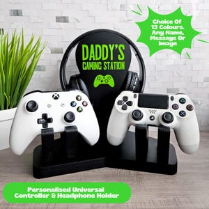 Puede incluir: Un soporte de auriculares para juegos negro con dos mandos de juego blancos. Los auriculares tienen una almohadilla negra y verde que dice "Daddy's Gaming Station".