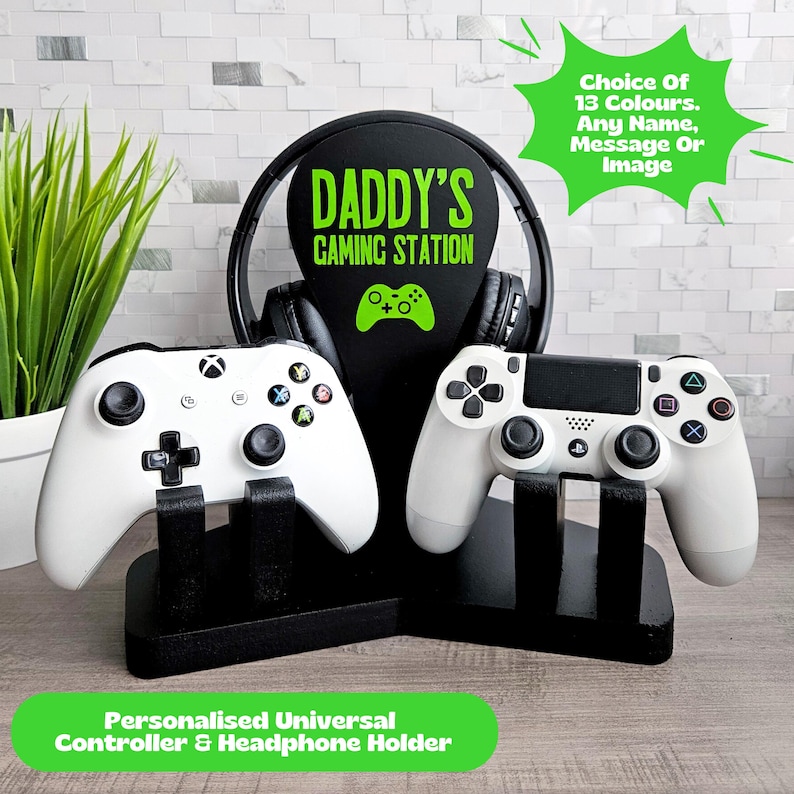 May include: A black gaming headset stand with two white game controllers. The headset has a black and green pad that says "Daddy's Gaming Station".