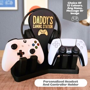 Może przedstawiać: Czarny stojak do gier z centralnym pionowym panelem z napisem DADDY'S GAMING STATION i ikoną kontrolera. Stojak mieści czarny zestaw słuchawkowy i dwa białe kontrolery. Tekst na obrazku brzmi Choice Of 13 Colours. Any Name, Message Or Image.