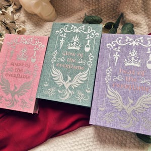 May include: Three hardcover books with textured fabric covers in pink, teal, and purple. Each cover features a white graphic of a crown, a dagger, a potion bottle, and a phoenix, with the titles Spark of the Everflame, Glow of the Everflame, and Heat of the Everflame.