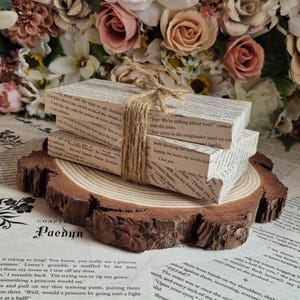 Puede incluir: Tres libros apilados, atados con cuerda, sobre una rodaja de madera rústica. Los libros están cubiertos de páginas de texto, con fragmentos de texto visibles. El fondo presenta un arreglo floral de rosas y otras flores.