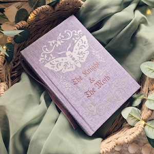May include: A lavender-colored book with the title "The Knight & The Moth" in burgundy script. The cover features a white moth illustration and decorative flourishes. The book rests in a woven basket on a green cloth.