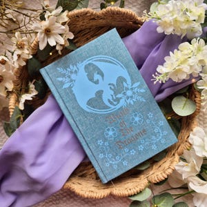 May include: A blue book with a dragon and floral design, titled "Wrath of the Dragons." The book is set in a woven basket with white flowers and a purple cloth.