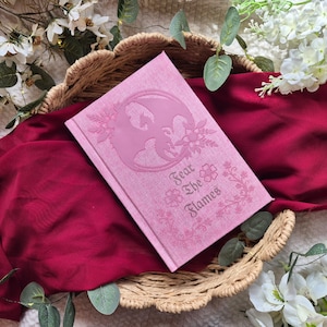 May include: A pink journal with the words "Fear the Flames" and a dragon illustration. The journal is in a woven basket on a red cloth, surrounded by white flowers and greenery.