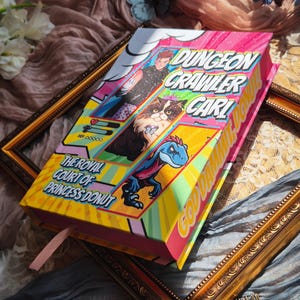 May include: A hardcover book titled Dungeon Crawler Carl: The Royal Court of Princess Donut with a vibrant, comic-book style cover featuring a person, a cat wearing a crown, and a blue dinosaur, with pink, yellow, and white graphic elements.