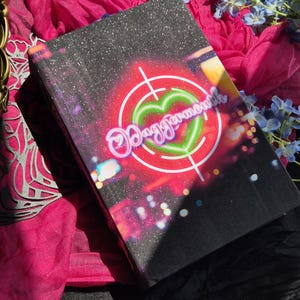 May include: A rectangular black box with a glittery finish featuring a graphic of a neon green heart inside a red target reticle. The text Daggermouth is written in a stylized script font across the center, surrounded by blurred colorful lights.