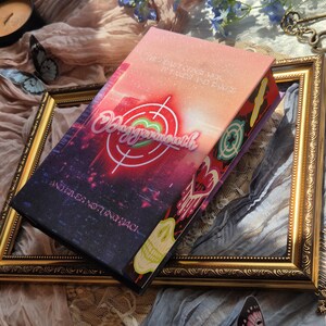 May include: A rectangular book with a gradient cover transitioning from orange to dark purple. The cover features a white target graphic, the text "Daggermouth", and the phrases "The heart consumes, it takes and takes" and "And gives nothing back."