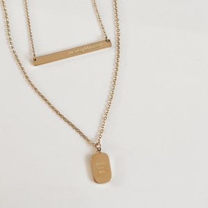 May include: Gold layered necklace with two pendants. The top pendant is a thin bar with the word "wingless" engraved on it. The bottom pendant is a rectangular tag with the words "with you" and "always" engraved on it.