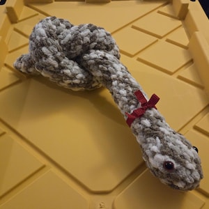 May include: A handmade plush snake toy in shades of gray and white, with a red ribbon tied around its neck. The snake has small, round, red eyes and is knotted in the middle. The toy is lying on a yellow surface.