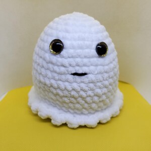 May include: White crocheted ghost with black eyes and a black mouth. The ghost is sitting on a yellow surface.