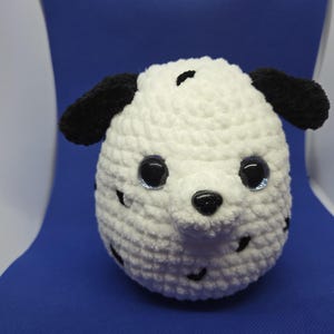 May include: A crocheted dog head with a white body and black spots. The dog has black ears, a black nose, and large, blue-rimmed eyes. The plushie is set against a blue background.