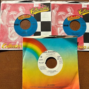 May include: Three 45 RPM vinyl records by Patsy Cline. Two records feature Collectables sleeves with red and white checkered patterns. The third record has a white label with black text and a rainbow graphic on the sleeve, titled Sweet Dreams.