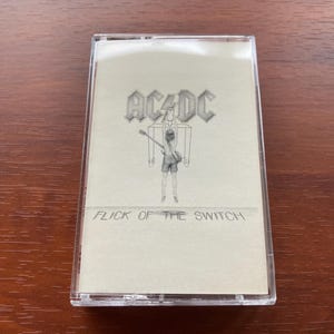 May include: AC/DC "Flick of the Switch" cassette tape. The cassette case is clear, and the album art features the band's logo and a figure playing a guitar. The text "Flick of the Switch" is below the image.
