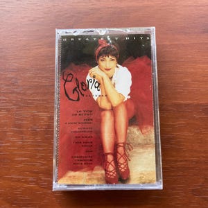 May include: A sealed cassette tape of Gloria Estefan's "Greatest Hits" album. The cover features a photo of Estefan in a white top and red skirt. The album title and song titles are printed in white and red.