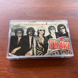 May include: A vintage cassette tape of The Traveling Wilburys album. The cover features a sepia-toned image of the band members in sunglasses and suits. The band's name is printed at the top, with the album title in a colorful, retro font.