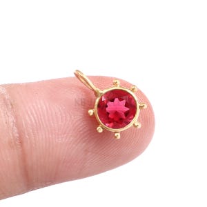 May include: A small gold pendant featuring a round, faceted, vibrant red gemstone set in a circular frame with six decorative bead accents around the perimeter and a small bail at the top for attachment to a chain or cord.