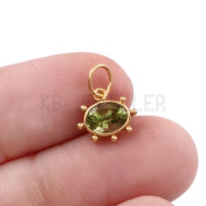 May include: A small gold charm featuring an oval-shaped, faceted olive green gemstone set in a metal frame with six decorative bead accents around the perimeter and a circular jump ring attached to the top for hanging purposes.