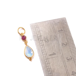 May include: A gold pendant featuring a pear-shaped, iridescent white gemstone and a small, round, red gemstone. The pendant is approximately 2.5 cm long. A ruler is next to the pendant.