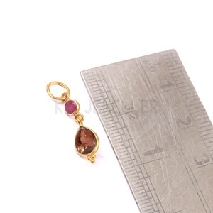 May include: A gold-colored pendant with a teardrop-shaped brown gemstone and a round, red gemstone. The pendant is suspended from a small loop. A ruler is in the background.