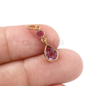 May include: A gold pendant featuring three gemstones. A small, round, red gemstone sits at the top. The middle section has a gold connector. The bottom gemstone is a large, teardrop-shaped, purple stone.
