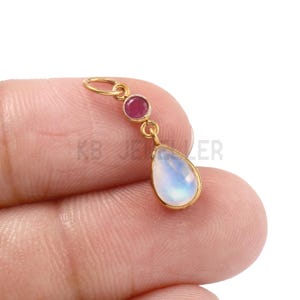 May include: A gold pendant featuring a teardrop-shaped moonstone and a small, round ruby. The pendant is suspended from a gold loop. The text "KB JEWELER" is visible in the background.