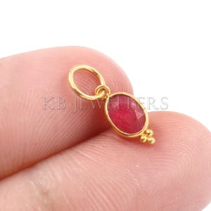 May include: Gold charm featuring a red oval gemstone and a small loop at the top. The charm has a gold bead detail at the bottom.