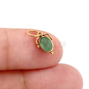 May include: A gold-colored pendant with an oval, green gemstone. The pendant has a loop at the top for a chain and decorative flourishes around the gemstone. The pendant is held between two fingers.