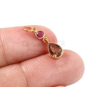 May include: A gold-colored pendant with a small, round, red gemstone and a larger, teardrop-shaped, brown gemstone. The pendant is held by a person's fingers.