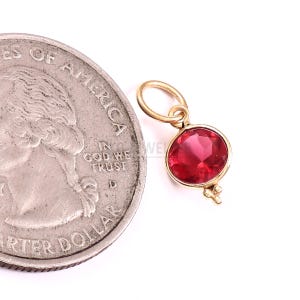 May include: A small gold pendant featuring a round, faceted red gemstone set in a gold bezel. The pendant includes a circular jump ring at the top and three tiny gold beads at the bottom, shown next to a United States quarter coin for scale.