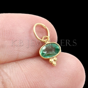 May include: A gold pendant featuring a green oval emerald gemstone with a gold bail and three small gold beads at the bottom.