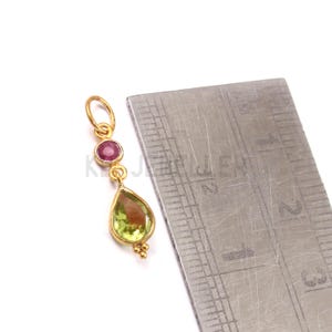 May include: A gold pendant featuring a teardrop-shaped green gemstone and a round red gemstone. The pendant is next to a ruler, showing measurements in centimetres.