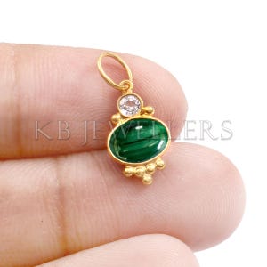 May include: A gold pendant featuring a vibrant green malachite stone in an oval shape, accented with a smaller, clear gemstone and gold detailing. The pendant has a loop for a chain. The text "KB JEWELLERS" is visible.
