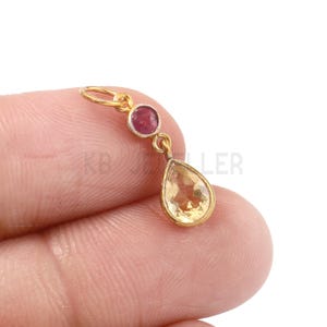 Könnte beinhalten: Ein goldfarbener Anhänger mit einem kleinen, runden, roten Edelstein oben und einem größeren, tropfenförmigen, gelben Edelstein darunter. Der Anhänger ist an einer kleinen Schlaufe befestigt.