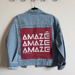 May include: Light blue denim jacket with a red patch on the back. The patch features the word "AMAZE" repeated three times in white, along with a camel graphic and the text "Roland and Percy".