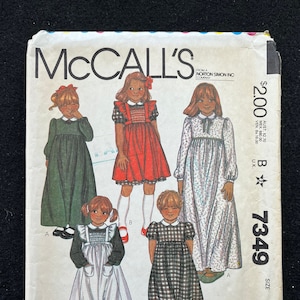 May include: McCall's sewing pattern number 7349 size B for a girl's dress. The pattern includes four different dress styles: a green dress with a white collar, a red dress with a white bib and white collar, a white dress with a green bib and white collar, and a plaid dress with a white collar.