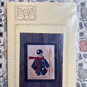 May include: A beige card with a brown wooden frame containing a cross-stitch image of a snowman holding a broom. The text "snowball" is below the snowman. The card also has a beige border with a dashed line pattern. The text "SNOWBALL SAMPLER #312" is at the bottom of the card.